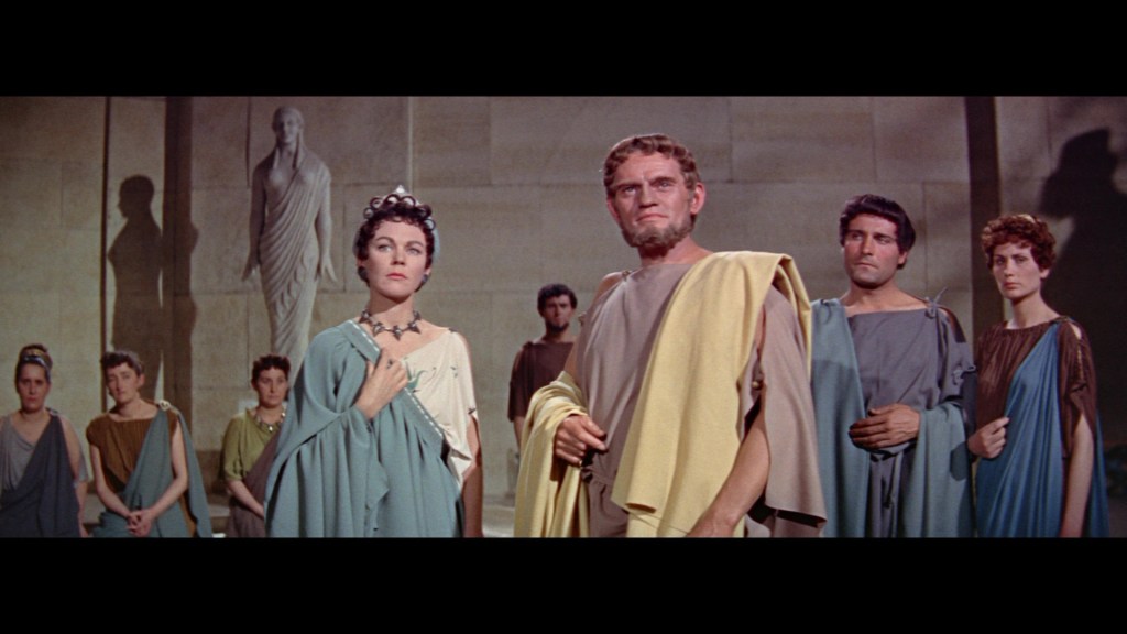 A group of people in ancient Roman clothing stands in a ceremonial setting, with a statue in the background. They exhibit expressions of seriousness and contemplation.