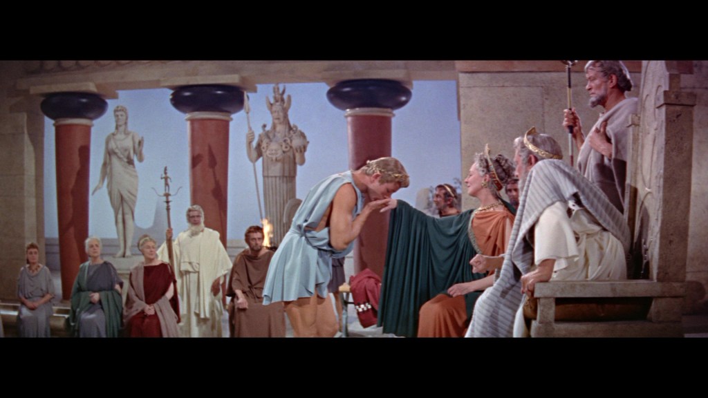 A scene depicting a dramatic moment in a classical setting, with a man in a blue tunic bowing to a seated woman in an ornate gown, surrounded by figures in ancient Roman attire and statues in the background.