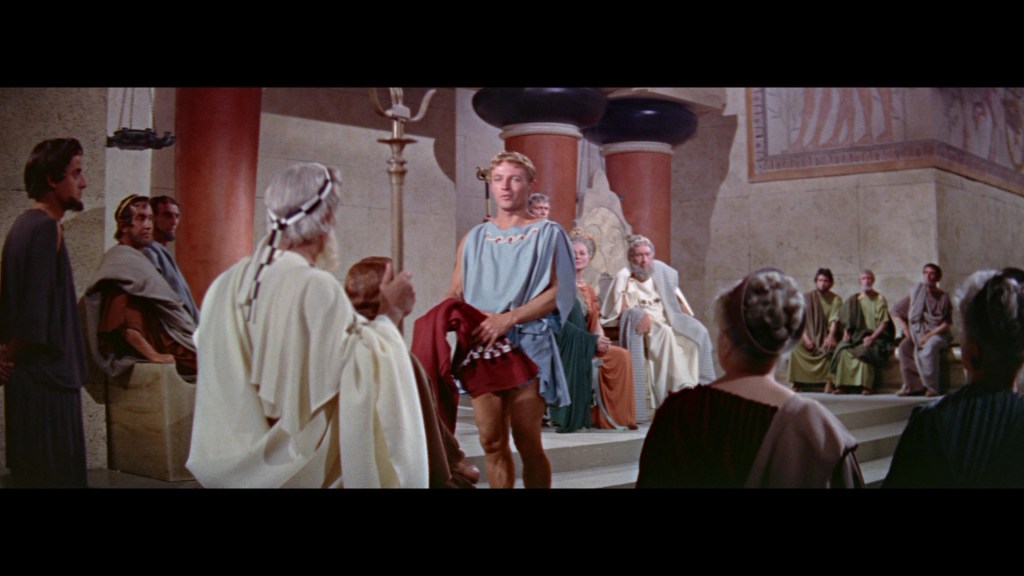 A scene depicting ancient Greek men in togas gathered in a large hall, with a central figure standing in a blue garment, seemingly addressing the seated audience.