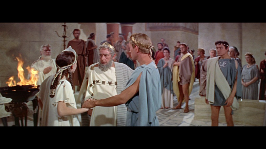A scene from a historical drama featuring a gathering of ancient Greek characters, with a central couple holding hands in conversation, surrounded by onlookers in ancient attire. In the background, a fire burns in a vessel.