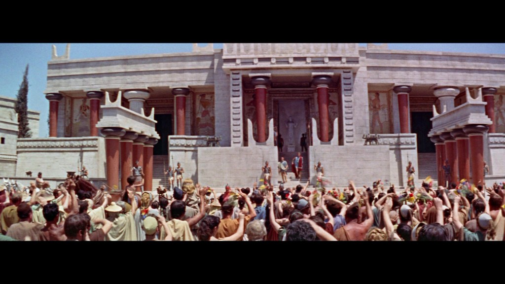 A large crowd of people in ancient attire gathers in front of an ornate stone building with red columns, cheering and holding flowers, set against a blue sky.