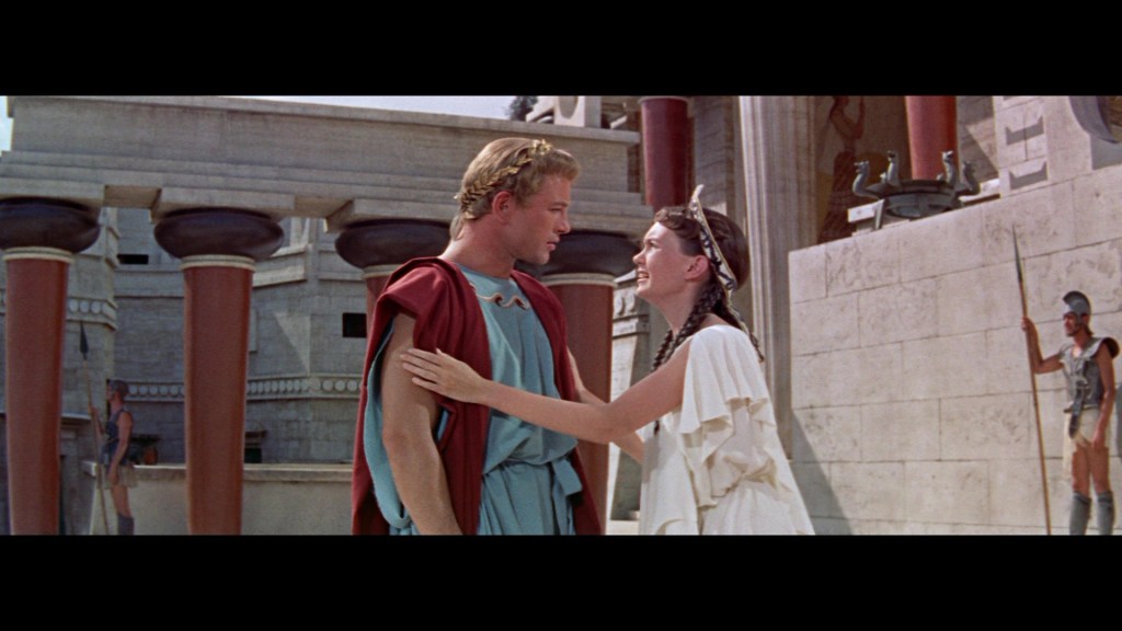 A scene featuring two characters in ancient Greek attire, a man in a blue tunic and red cape, and a woman in a white dress with a headpiece, sharing an intimate moment in a historical setting with columns and a stone backdrop.