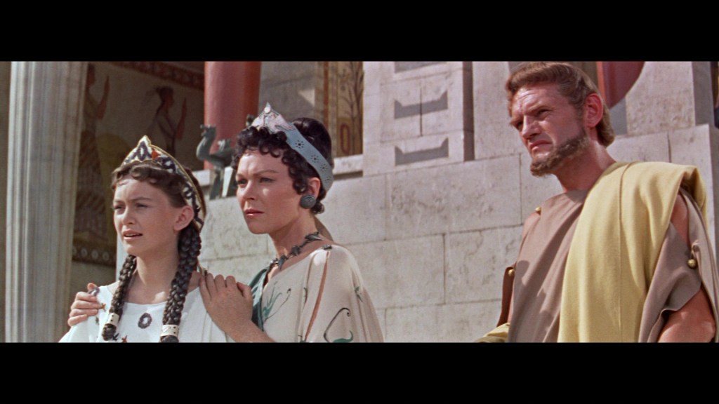 Three characters in ancient attire stand together, with a backdrop of classical architecture and mural artwork. One woman, dressed in white with a crown, appears concerned, while another girl beside her holds her arm. A man in a toga stands on the right, looking serious.
