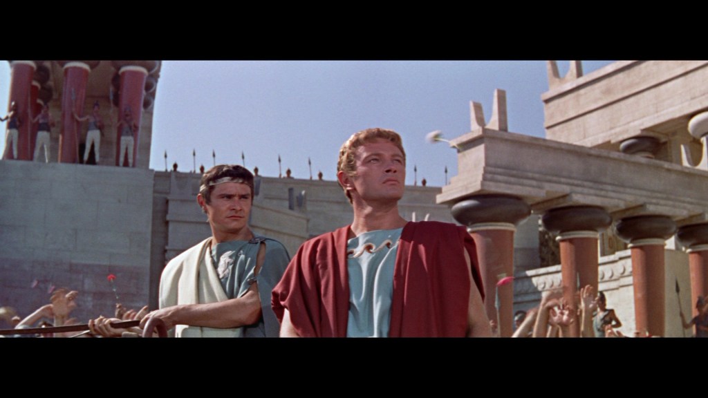 Two men in ancient Roman attire standing in a public square, with a crowd and classical architecture in the background.