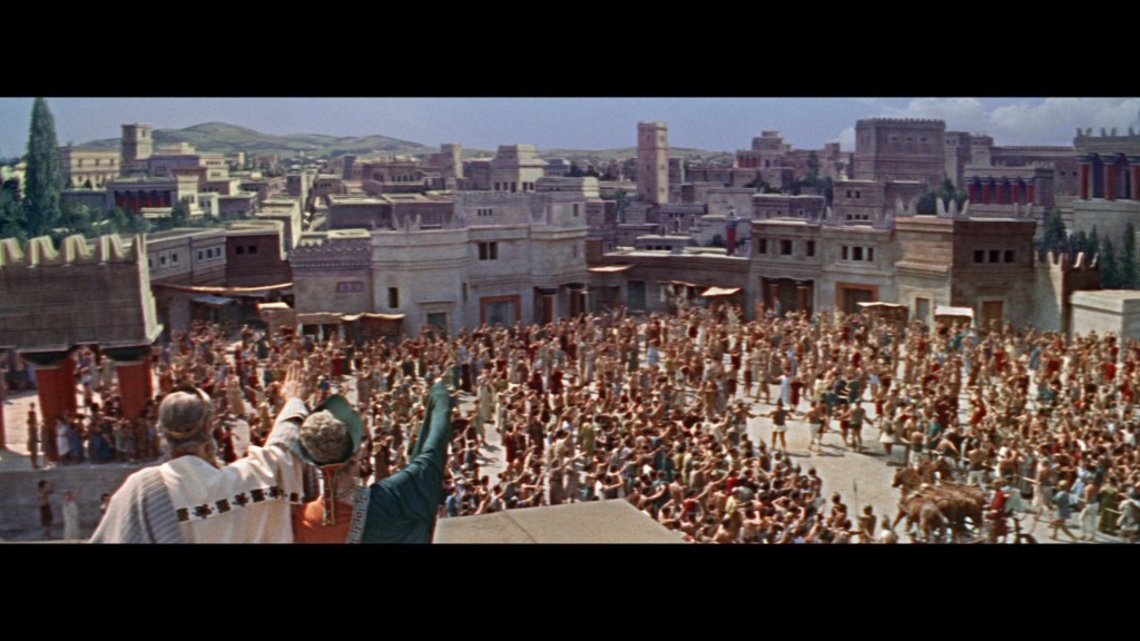 A large crowd of people gathered in an ancient city setting, with prominent buildings in the background. Two figures on a raised platform are addressing the crowd, who appear enthusiastic and engaged.