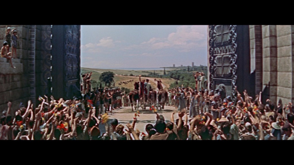 A large crowd gathered outdoors, cheering enthusiastically as figures on chariots enter through massive stone gates, under a bright blue sky with scattered clouds.
