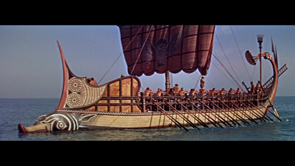 A large ancient ship with a patterned sail and decorative prow, sailing on calm blue waters, with numerous people on board.