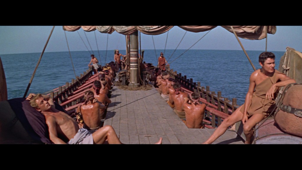 A view of a ship at sea with several men sitting and lounging on the deck, some engaged in labor. The scene captures a sunny day with clear skies and calm waters.