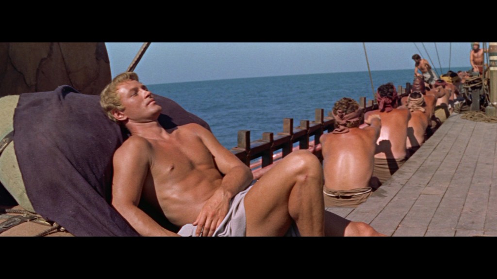 A shirtless man lounging on a ship deck, with ocean and sky in the background, while several other shirtless figures are seen in the distance.