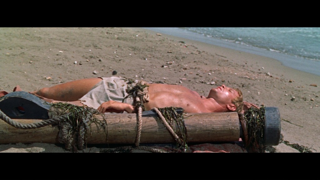 A muscular man lying on a sandy beach, bound to a large log with ropes and seaweed, against a backdrop of ocean waves.