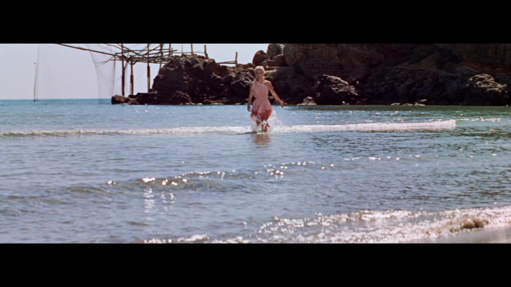 A woman in a pink dress wades through shallow water at a beach, with rocky cliffs and a fishing structure in the background under a clear blue sky.