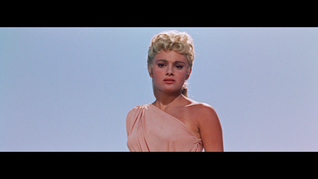 A young woman with curly blonde hair, wearing a one-shoulder dress, stands against a clear blue sky, looking solemn.