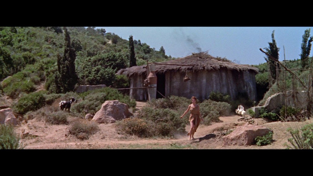 A person running in a natural landscape near a grass-covered hut, surrounded by greenery and rocks.