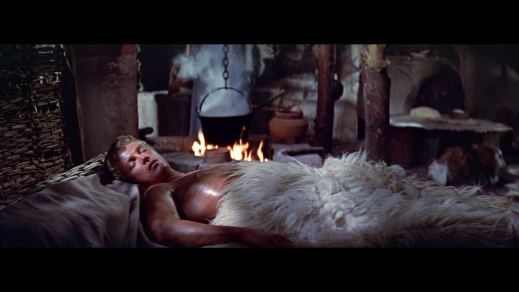 A young man lying on a bed covered with a fur blanket, in a dimly lit room with a fire in the background and various rustic items around.