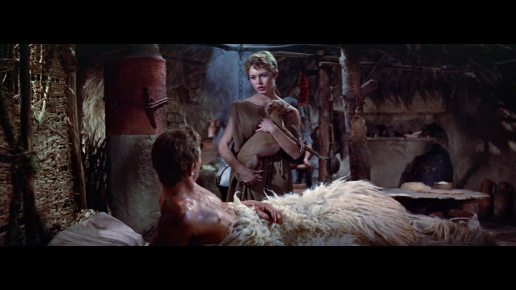 A woman holding a small animal stands over a man lying on a fur-covered bed in a rustic interior, surrounded by natural materials.