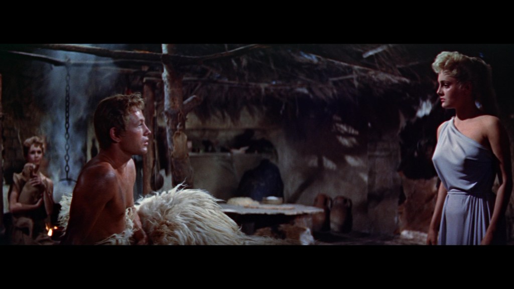 A scene inside a rustic dwelling with three characters: a shirtless man, a woman in a gray dress, and another woman in the background holding a small child, evoking a dramatic atmosphere.