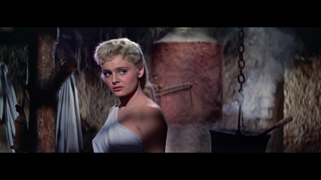 A blonde woman wearing a white garment stands in a dimly lit interior, looking back over her shoulder.