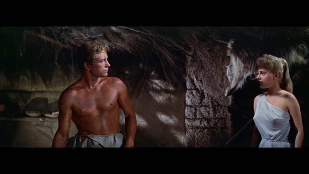A shirtless man stands facing a woman in a white dress, both looking at each other in a rustic setting with stone and thatched elements.