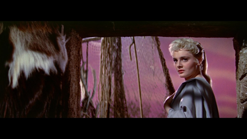 A woman with blonde hair and a gray cape appears in a doorway, gazing thoughtfully into the distance with a pink and orange sky in the background.
