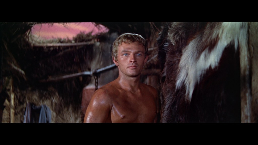 A muscular man with short blonde hair stands shirtless inside a rustic shelter, with a blurred background suggesting a sunset.
