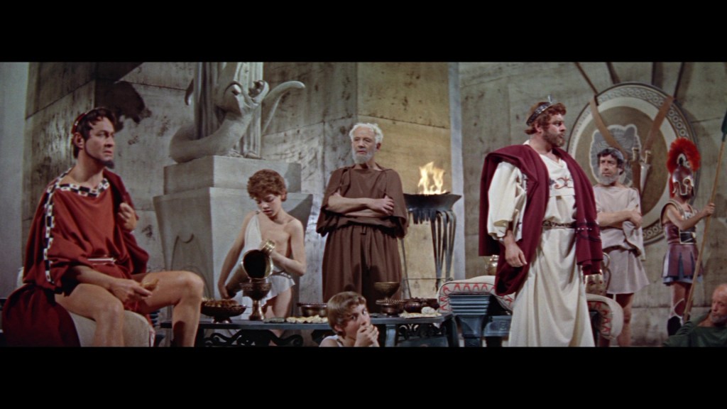 Scene depicting a historical gathering with several characters in ancient Roman attire, seated and standing around a table filled with dishes and artifacts, featuring a backdrop of stone walls and a torch.
