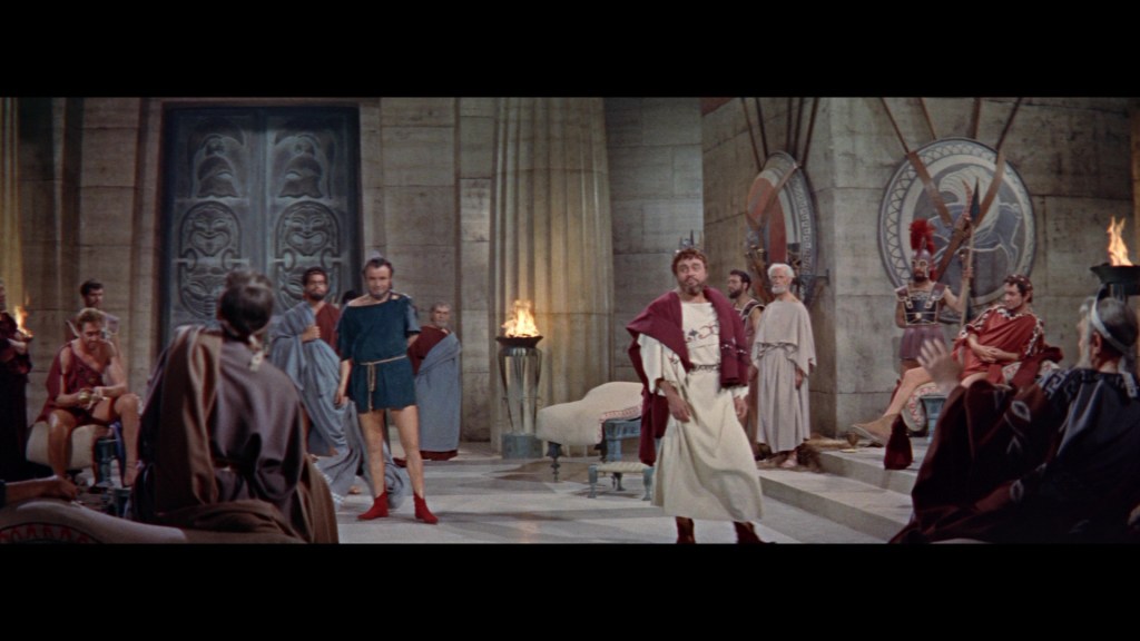A dramatic scene set in an ancient Roman court, with a group of men dressed in togas and traditional Roman attire, engaged in discussion. The main figure stands at the center, surrounded by others, while columns and ancient artwork decorate the background.