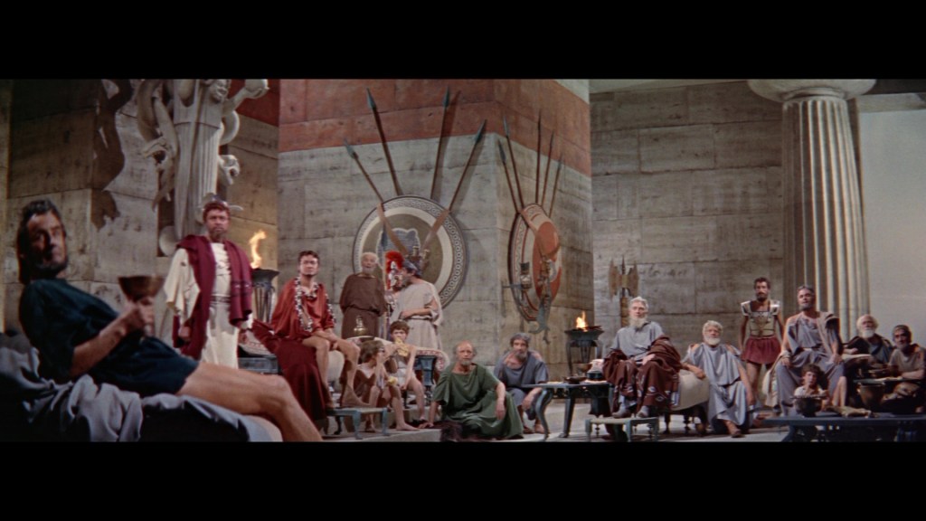 A group of ancient Roman figures seated in a grand hall, adorned with classical architecture, sculptures, and decorative shields. The men are dressed in togas and robes, engaged in conversation or looking towards the viewer.