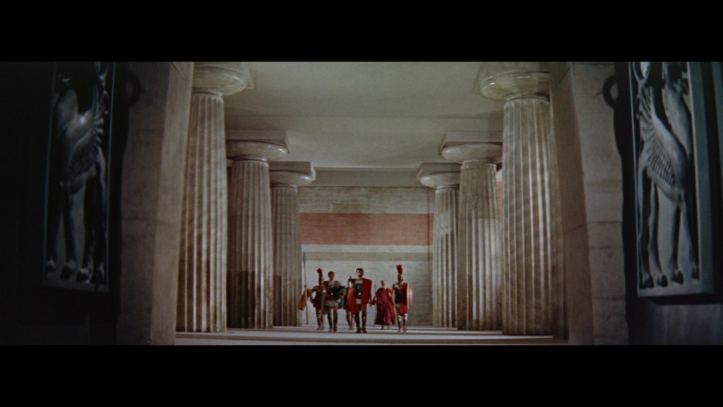 A group of figures dressed in ancient Roman attire walks through a grand corridor lined with tall columns and artwork on the walls.
