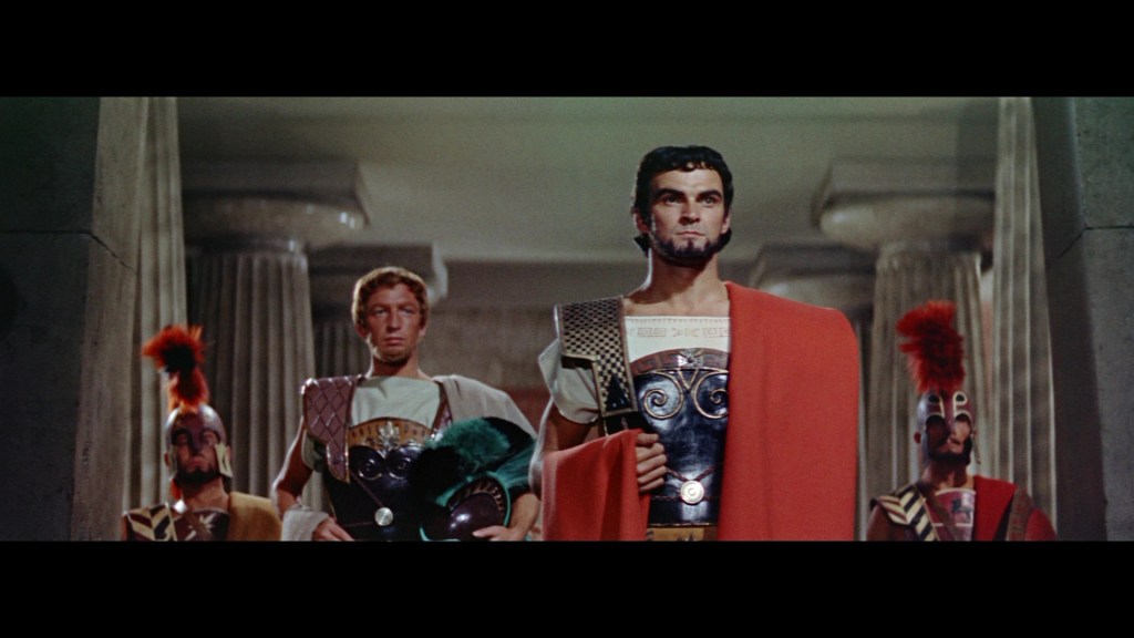 A group of four men in ancient Roman clothing, with one man in the front displaying a strong stance, wearing an elaborate tunic and a red cape, flanked by three others in armor, all set against a backdrop of columns.