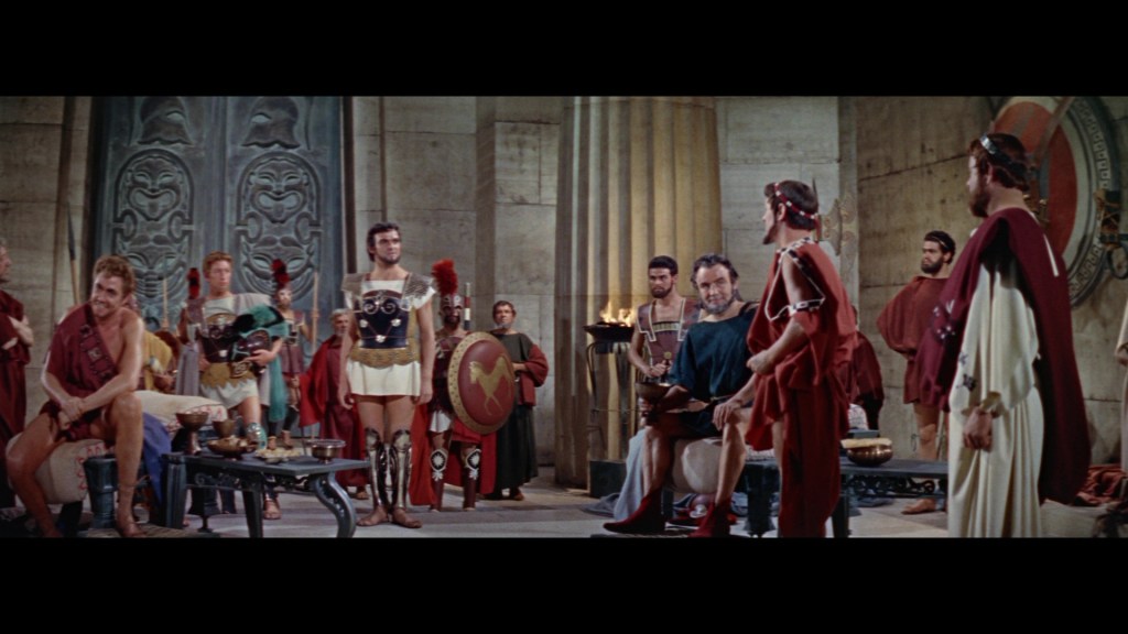 A scene depicting a group of ancient Roman men in traditional clothing, gathered in a grand chamber. Some men are seated while others stand, wearing armor and togas. The background features large stone columns and an ornate design on the wall.
