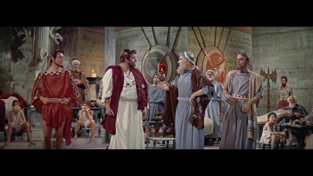 A scene depicting a group of ancient Greek men in traditional attire, engaged in conversation in a stylized hall, with several observers seated around them.