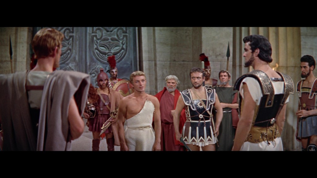 A dramatic scene featuring a group of ancient Greek soldiers and leaders, engaged in a tense discussion indoors, with ornate stonework and pillars in the background.