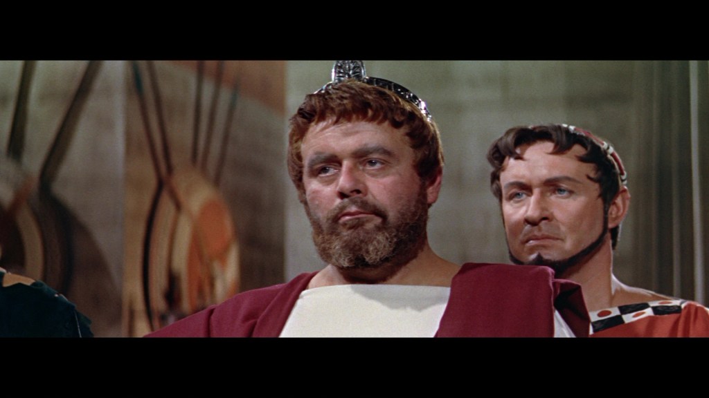 A scene depicting two Roman men, one with a beard and wearing a toga, looking serious, while a second man stands behind him, observing with a concerned expression.