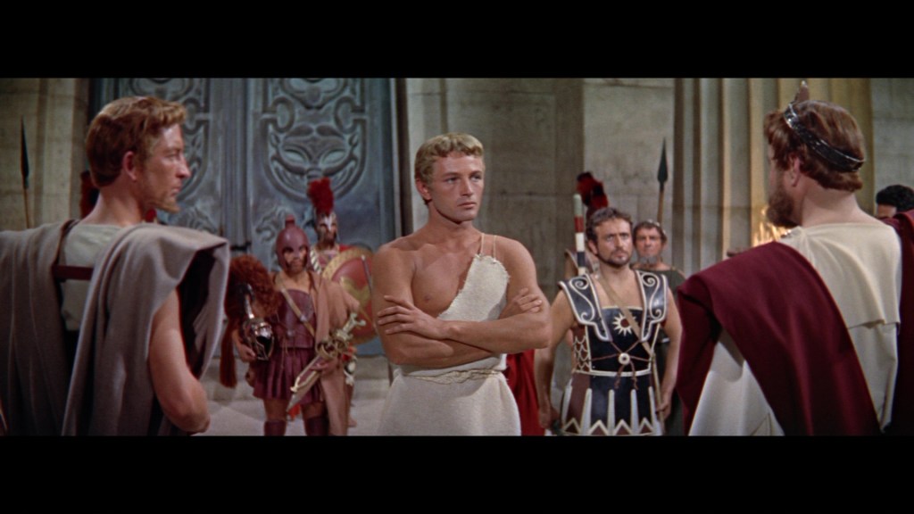 A scene depicting ancient Greek characters in a dramatic confrontation, featuring a muscular man in a white tunic standing at the center, flanked by others in historical attire, set against a grand stone backdrop.
