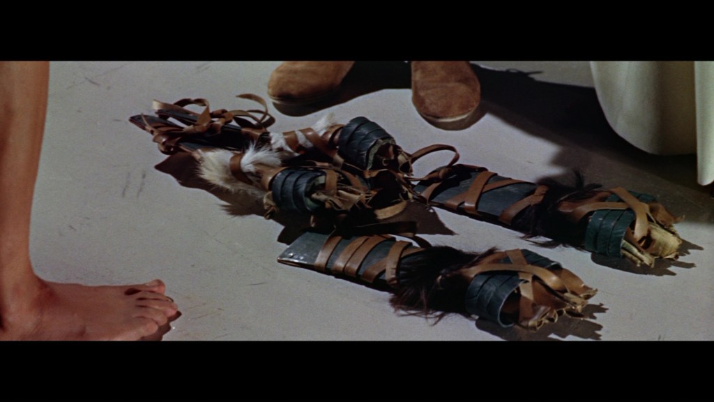 A close-up view of traditional footwear made of leather and various straps, placed on a smooth floor. A bare foot is visible in the foreground, along with a person's brown shoe in the background.