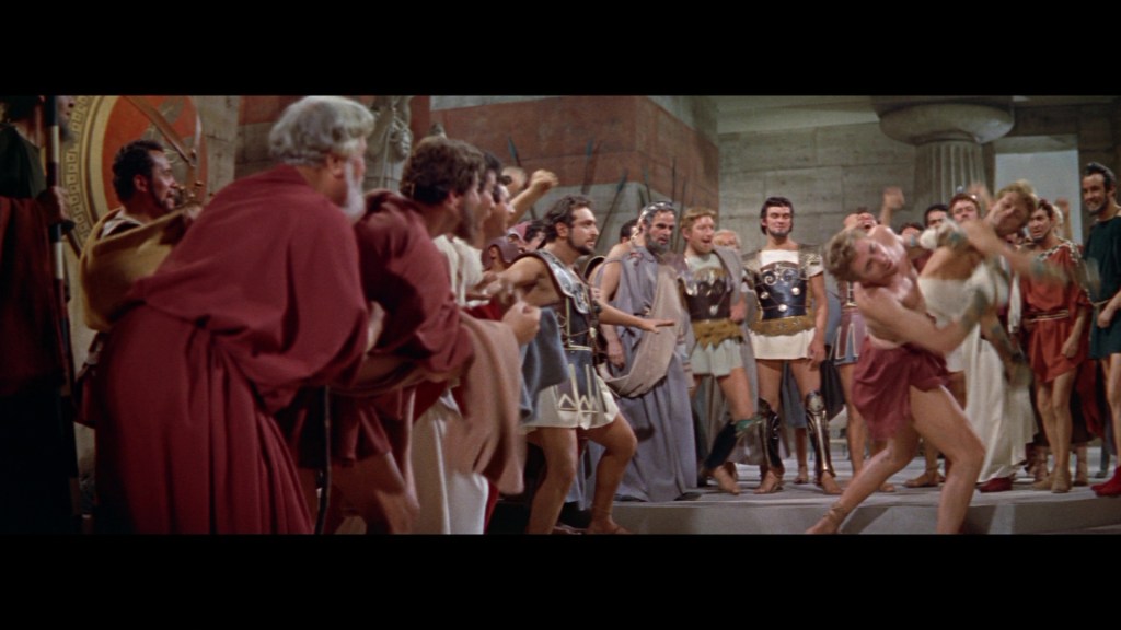 A lively scene from an ancient Roman gathering, featuring a crowd of men and women in period costumes, some cheering and others performing, set in a grand hall with stone architecture.