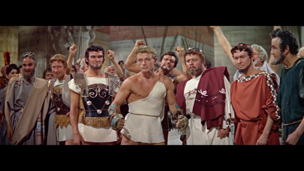 A group of ancient Roman men, including a muscular fighter in a gladiator outfit, stands together in a scene depicting a historical or theatrical moment, with expressions of determination and excitement.