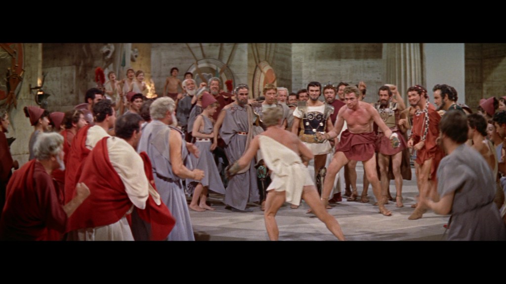 A lively scene from a historical drama featuring a large group of characters in ancient Roman attire, gathered in an arena setting, with intense focus on two individuals engaged in a competitive display.