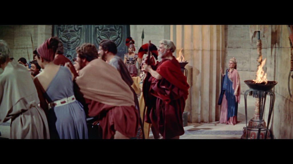A scene set in ancient times, depicting a crowd of people dressed in traditional Roman attire, with a focus on a figure in a red robe holding a staff, and a woman in the background wearing a blue dress.