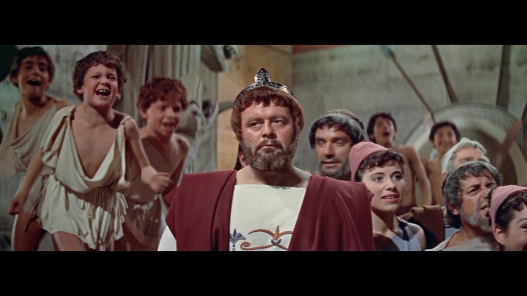 A scene portraying a group of children and adults in historical attire, with a central figure wearing a crown and looking intense, surrounded by lively reactions from the children and other characters.