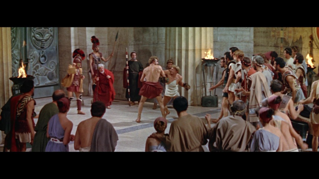 A historical scene depicting a gladiatorial match in an ancient Roman arena, with spectators cheering and various characters in traditional costumes. The focus is on two gladiators preparing to fight, while soldiers and onlookers surround them.