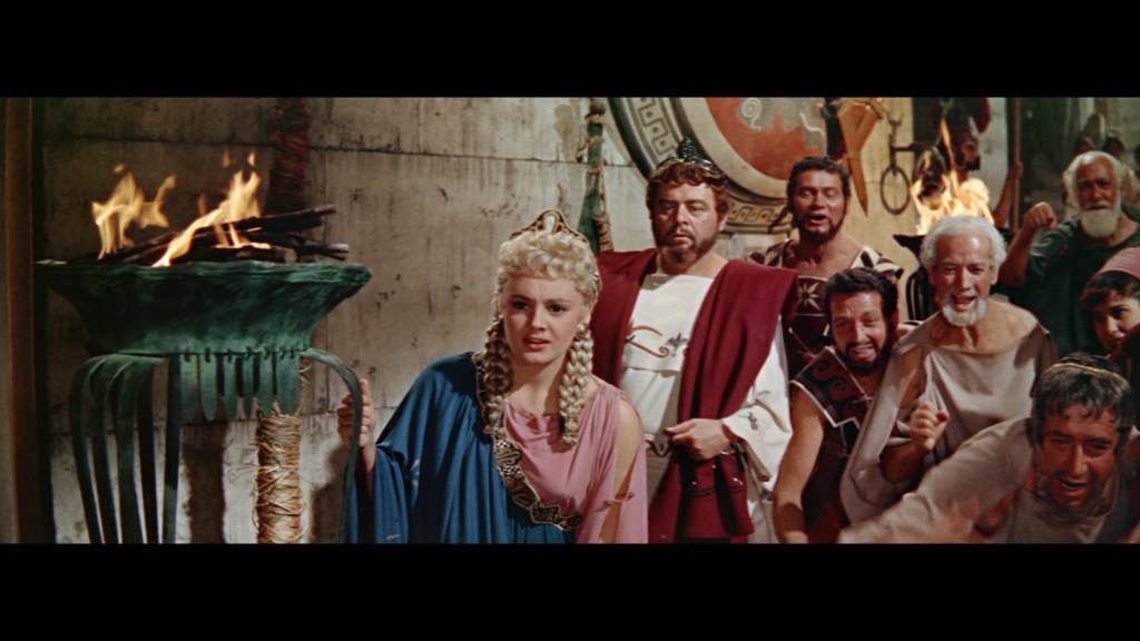 A scene depicting a group of ancient Roman characters, with a woman in a blue and pink dress and several men in togas, gathered around a lit torch in a dramatic setting.