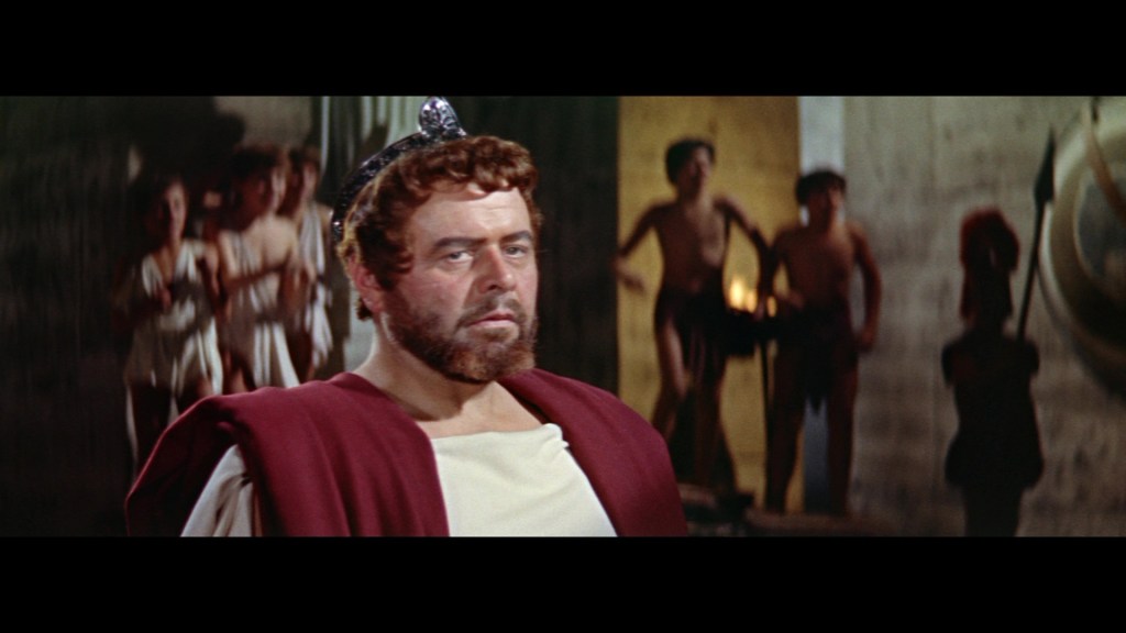 A historical character in a toga and crown, with a serious expression, standing in a dimly lit setting with people in ancient clothing in the background.