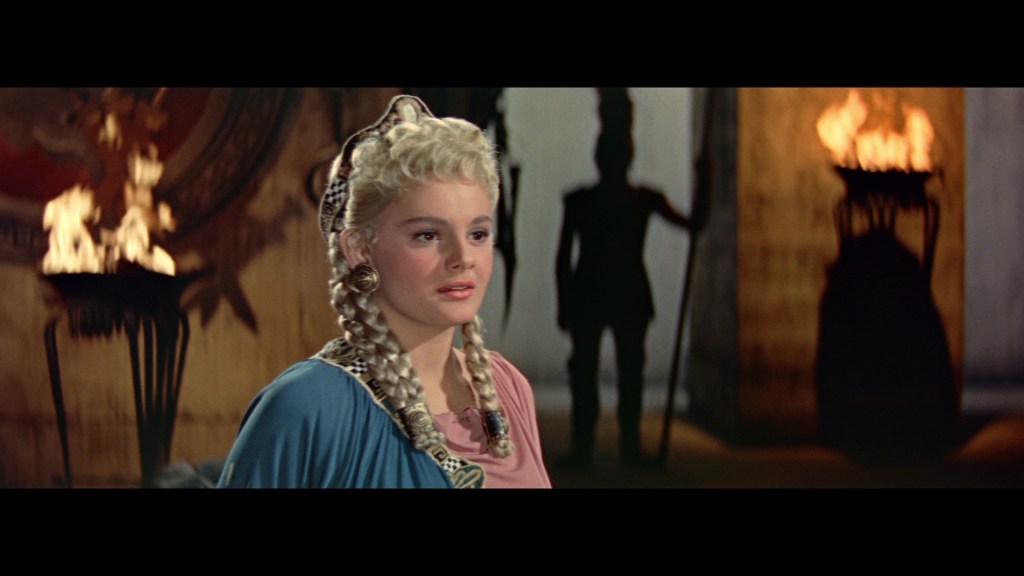 A woman with blonde hair styled in braids, wearing a blue and pink dress, stands in a dimly lit room with flames in the background casting shadows.