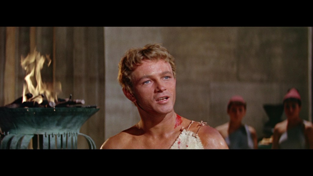 A young man with tousled hair and a determined expression stands in front of a burning torch, wearing a partially torn garment, with a few visible injuries on his shoulder, while two figures in the background observe.