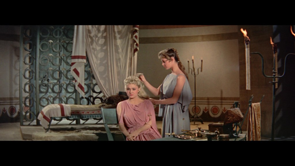 A scene depicting two women in an ancient setting. One woman with blonde hair is seated, while the other, dressed in a light blue dress, is styling her hair. The background features decorative elements and candlelit ambiance.