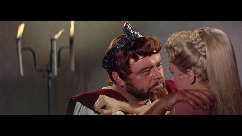 A man wearing a crown gazes intensely at a woman while embracing her, with candles in the background.