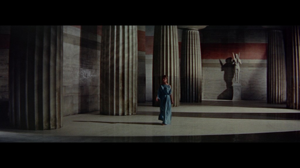 A woman in a long blue dress walks through a large, ancient-inspired hall with tall columns and a statue in the background.