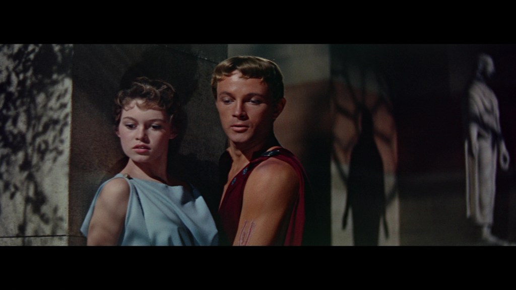 A dramatic moment between a man and woman, with the woman wearing a light blue dress and the man in a sleeveless red garment, both standing close together against a background with shadows and a statue.