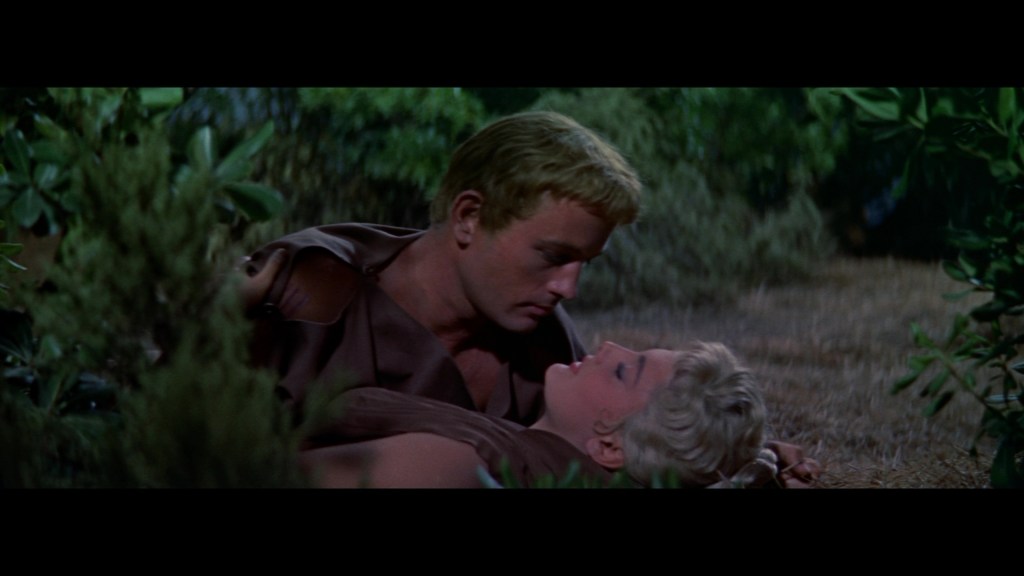 A close-up scene of a man and woman lying on the ground amidst greenery, gazing into each other's eyes with an intimate expression.